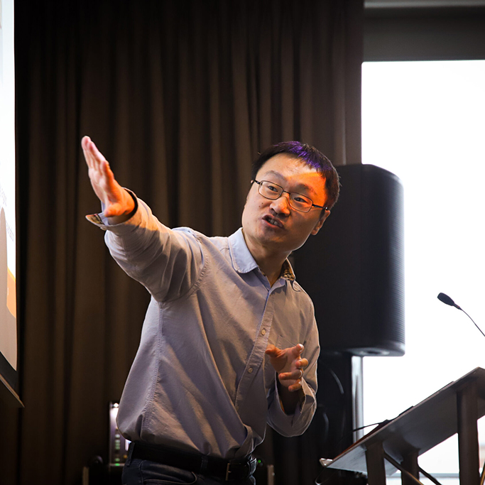 Professor He Wang is pictured mid-presentation. The projector screen light reflects on his face.